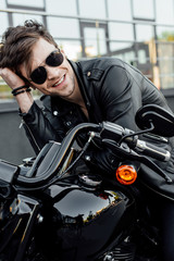 Fototapeta na wymiar handsome young motorcyclist in sunglasses holding head with hand, smiling and looking away while sitting on motorcycle