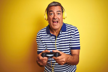 Middle age man playing video game using headphones over isolated yellow background scared in shock with a surprise face, afraid and excited with fear expression © Krakenimages.com