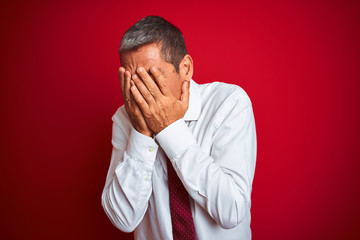 Handsome middle age businessman standing over isolated red background with sad expression covering face with hands while crying. Depression concept.
