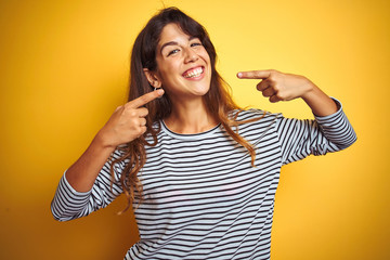 Young beautiful woman wearing stripes t-shirt standing over yelllow isolated background smiling cheerful showing and pointing with fingers teeth and mouth. Dental health concept.