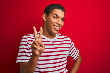 Young handsome arab man wearing striped t-shirt over isolated red background smiling looking to the camera showing fingers doing victory sign. Number two.