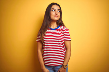 Young beautiful woman wearing striped t-shirt standing over isolated yellow background smiling looking to the side and staring away thinking.