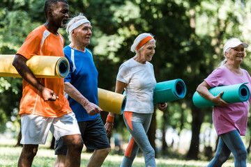 selective focus of positive retired and multiethnic pensioners holding fitness mats while walking...