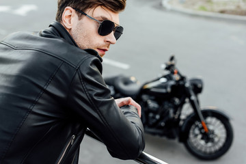 Fototapeta na wymiar selective focus of young man in black leather jacket standing near metal fence with motorcycle on background