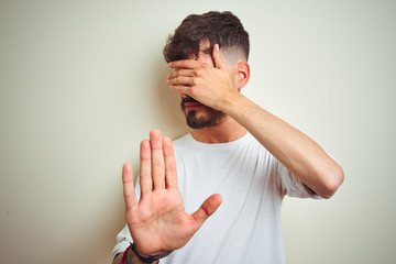Young man with tattoo wearing t-shirt standing over isolated white background covering eyes with hands and doing stop gesture with sad and fear expression. Embarrassed and negative concept.