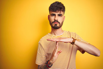 Young man with tattoo wearing striped t-shirt standing over isolated yellow background Doing time out gesture with hands, frustrated and serious face