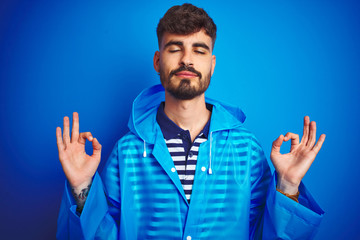 Young handsome man wearing rain coat standing over isolated blue background relax and smiling with eyes closed doing meditation gesture with fingers. Yoga concept.