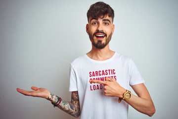 Young man with tattoo wearing fanny t-shirt standing over isolated white background amazed and...