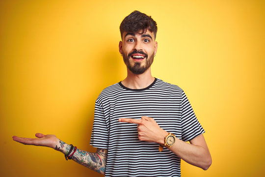 Young man with tattoo wearing striped t-shirt standing over isolated yellow background amazed and smiling to the camera while presenting with hand and pointing with finger.