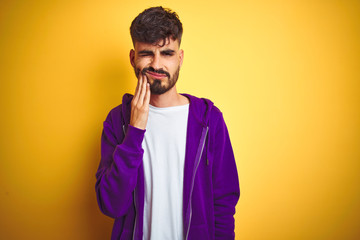 Young man with tattoo wearing sport purple sweatshirt over isolated yellow background touching mouth with hand with painful expression because of toothache or dental illness on teeth. Dentist concept.