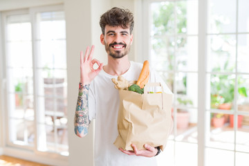 Young man holding paper bag of fresh groceries from the supermarket doing ok sign with fingers, excellent symbol