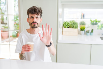 Young man drinking a coffee in a paper cup in the morning with open hand doing stop sign with serious and confident expression, defense gesture