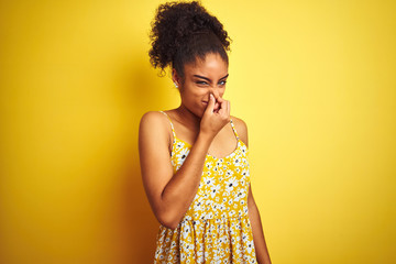 African american woman wearing casual floral dress standing over isolated yellow background smelling something stinky and disgusting, intolerable smell, holding breath with fingers on nose. Bad smells - obrazy, fototapety, plakaty