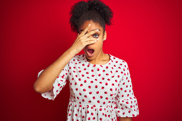Fototapeta na wymiar African american woman wearing fashion white dress standing over isolated red background peeking in shock covering face and eyes with hand, looking through fingers with embarrassed expression.