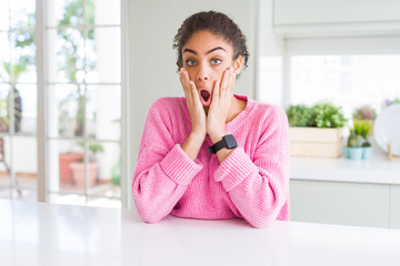 Fototapeta na wymiar Beautiful african american woman with afro hair wearing casual pink sweater afraid and shocked, surprise and amazed expression with hands on face