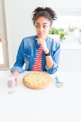 Young african american woman eating homemade cheese pizza serious face thinking about question, very confused idea