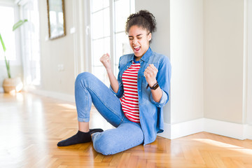 Beautiful young african american woman with afro hair sitting on the floor very happy and excited doing winner gesture with arms raised, smiling and screaming for success. Celebration concept.