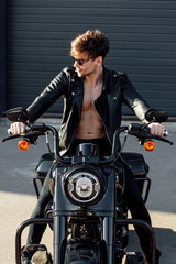 handsome motorcyclist with muscular naked torso in leather jacket sitting on motorcycle, looking...