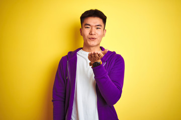 Fototapeta premium Young asian chinese man wearing purple sweatshirt standing over isolated yellow background looking at the camera blowing a kiss with hand on air being lovely and sexy. Love expression.