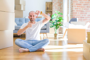 Young bald man sitting on the floor around cardboard boxes moving to a new home smiling making frame with hands and fingers with happy face. Creativity and photography concept.