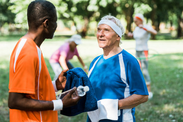 Fototapeta premium selective focus of happy and sportive senior man holding bottle with water and looking at african american man