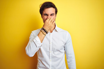 Young handsome businessman wearing elegant shirt standing over isolated yellow background smelling something stinky and disgusting, intolerable smell, holding breath with fingers on nose. Bad smells 