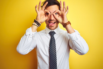 Young handsome businessman wearing elegant shirt and tie over isolated yellow background doing ok gesture like binoculars sticking tongue out, eyes looking through fingers. Crazy expression.