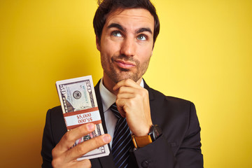Young handsome businessman wearing suit holding dollars over isolated yellow background serious...