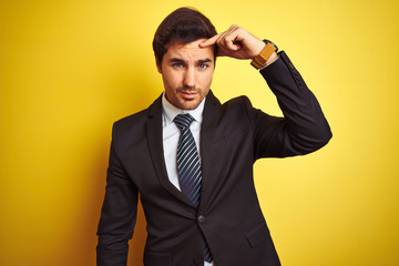 Young handsome businessman wearing suit and tie standing over isolated yellow background pointing unhappy to pimple on forehead, ugly infection of blackhead. Acne and skin problem
