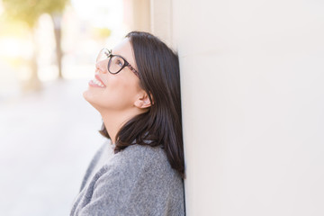 Beautiful young woman wearing glasses smiling cheerful leaning on wall, casual pretty girl at the town