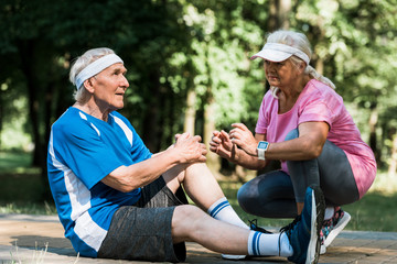 retired man touching knee while sitting on walkway near senior woman in cap