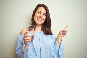 Young beautiful woman drinking a glass of fresh water over isolated background very happy pointing with hand and finger to the side