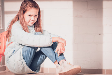 Good girl with flowing hair in blue down jacket sits and hugs her knees with her hands against background of white brick wall.