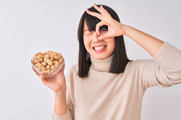 Young beautiful Chinese woman holding bowl with peanuts over isolated white background with happy face smiling doing ok sign with hand on eye looking through fingers