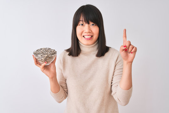 Beautiful chinese woman holding bowl with sunflowers seeds over isolated white background surprised with an idea or question pointing finger with happy face, number one