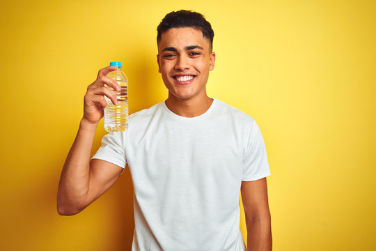 Young brazilian man holding bottle of water standing over isolated yellow background with a happy face standing and smiling with a confident smile showing teeth