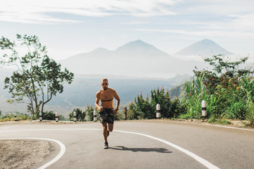 Man running topless in uphill on the asphalt road in hot summer weather. Panoramic mountain view on...