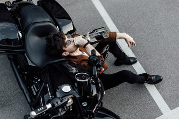 top view of young motorcyclist sitting on ground, leaning on motorcycle and drinking alcohol