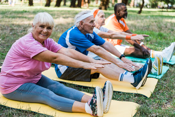 selective focus of happy senior woman in sportswear exercising near multicultural pensioners on fitness mats