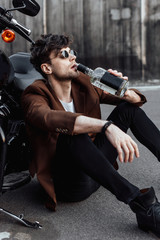Fototapeta na wymiar young man drinking alcohol while sitting on ground near motorcycle