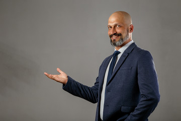 Cute young bald bearded businessman in a classic suit with an open palm in which to place your subject. Gray background with copy space