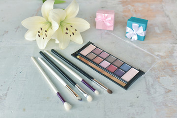 Make up brushes, applicator, eye shadows and a blooming flowers on a white wooden table.