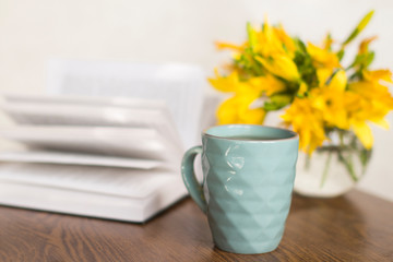 blue coffee mug, yellow flowers in transparent bowl and a book on a wooden table