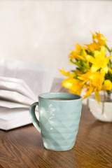 blue coffee mug, yellow flowers in transparent bowl and a book on a wooden table