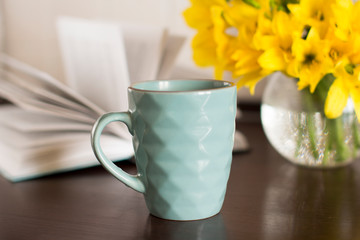 blue coffee mug, yellow flowers in transparent bowl and a book on a wooden table