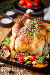 Baked whole chicken with vegetables and herbs close-up on a plate on a table.