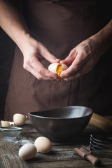 Professional chef hands are breaking an egg into bowl to make dough on wooden table, over dark background