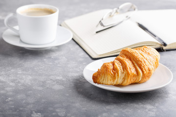 Morning cup of coffee and croissant for breakfast. Day planning, empty notebook, pen and glasses on gray table.