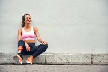 Simply happy Young smiling woman with leg prosthesis in sports clothing and headphones listening music after her workout while sitting outdoors