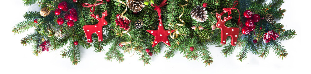 Festive Christmas border, isolated on white background. Fir green branches are decorated with red stars, berries, fir cones and red toy deer, close-up.